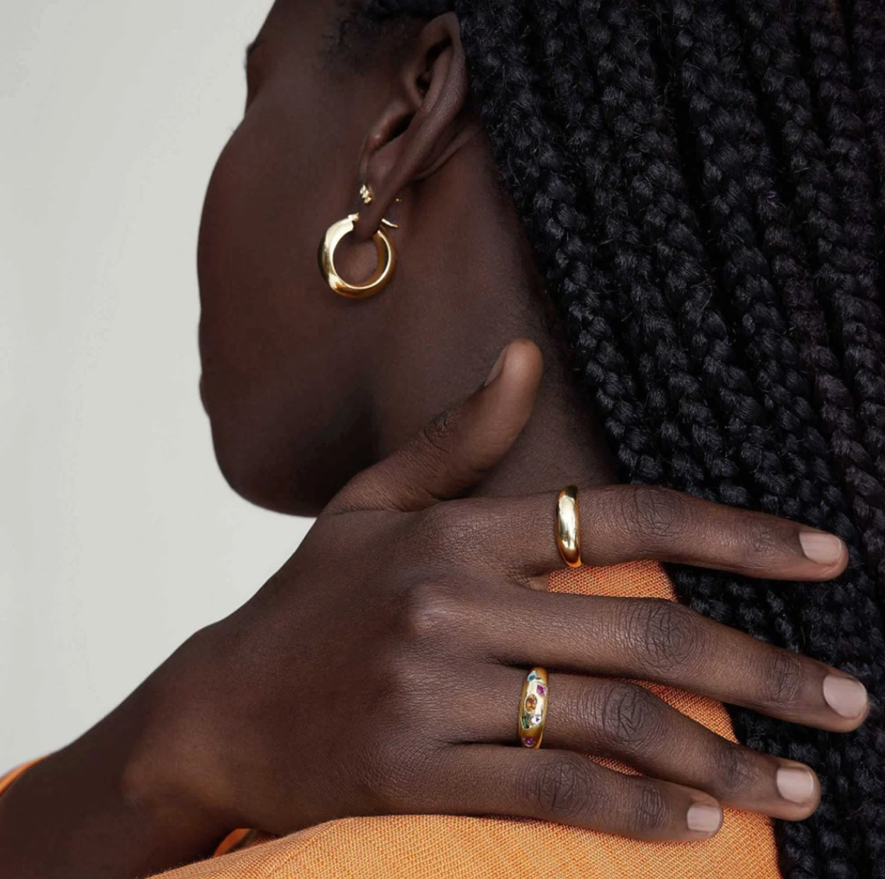Close-up of a person wearing gold hoop earrings and rings on a neutral background