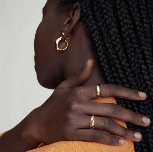 Close-up of a person wearing gold hoop earrings and rings on a neutral background